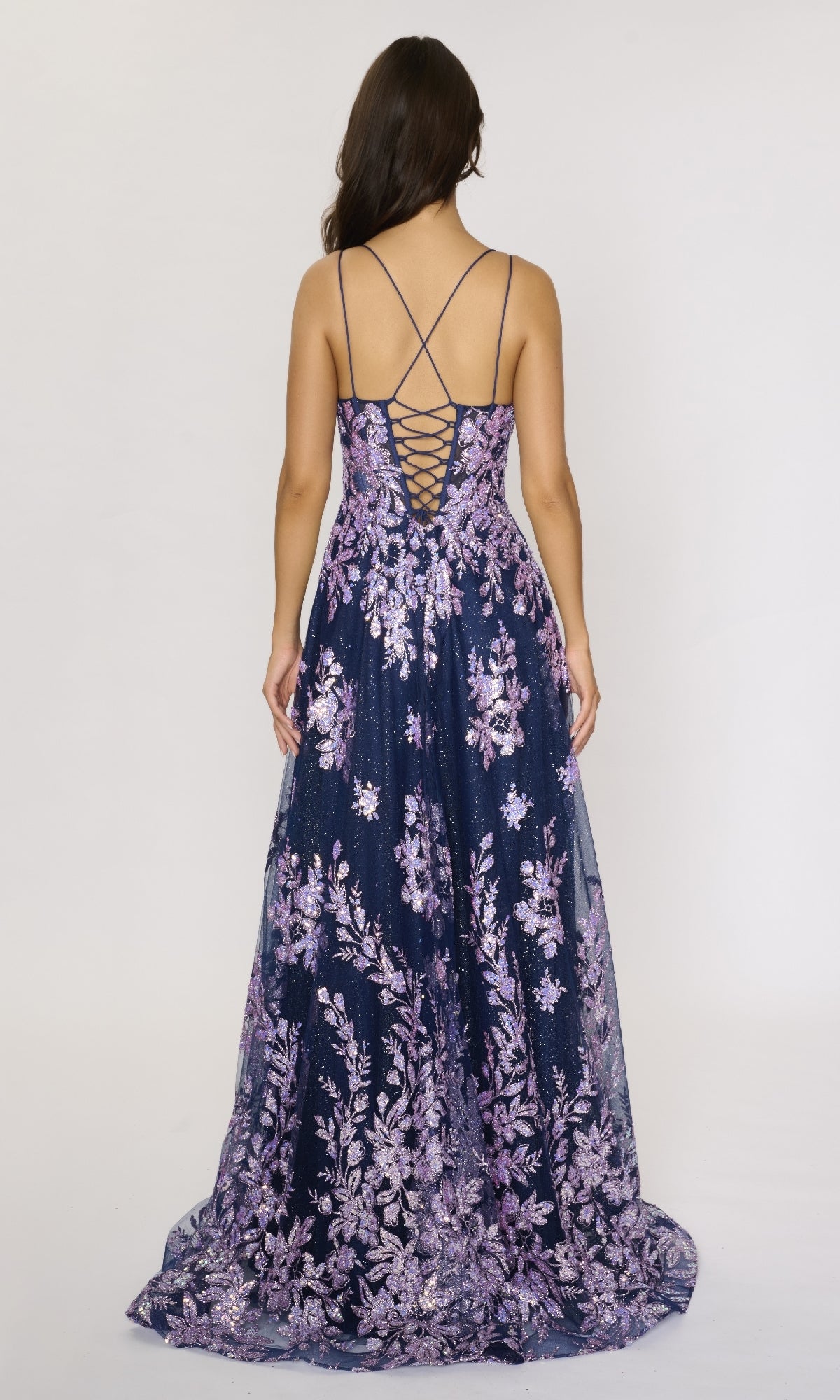 A woman stands facing away in the Nina Canacci 1589 long formal dress, featuring dark blue and purple glitter, thin straps, an embellished bodice, and a lace-up back.